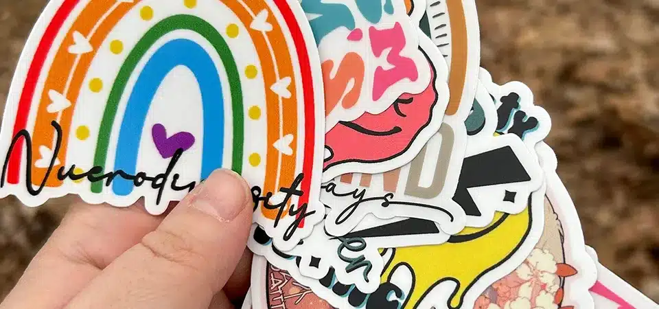 A collection of various waterproof stickers: a white die-cut, a clear sticker on glass, and a holographic sticker