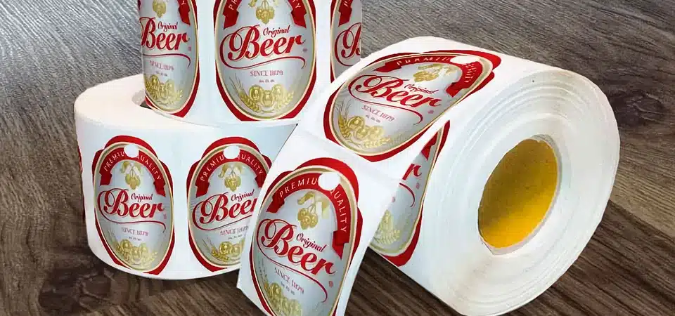 A side-by-side comparison showing a thick vinyl sticker peeling off a curved bottle, while a thin waterproof label stays flat